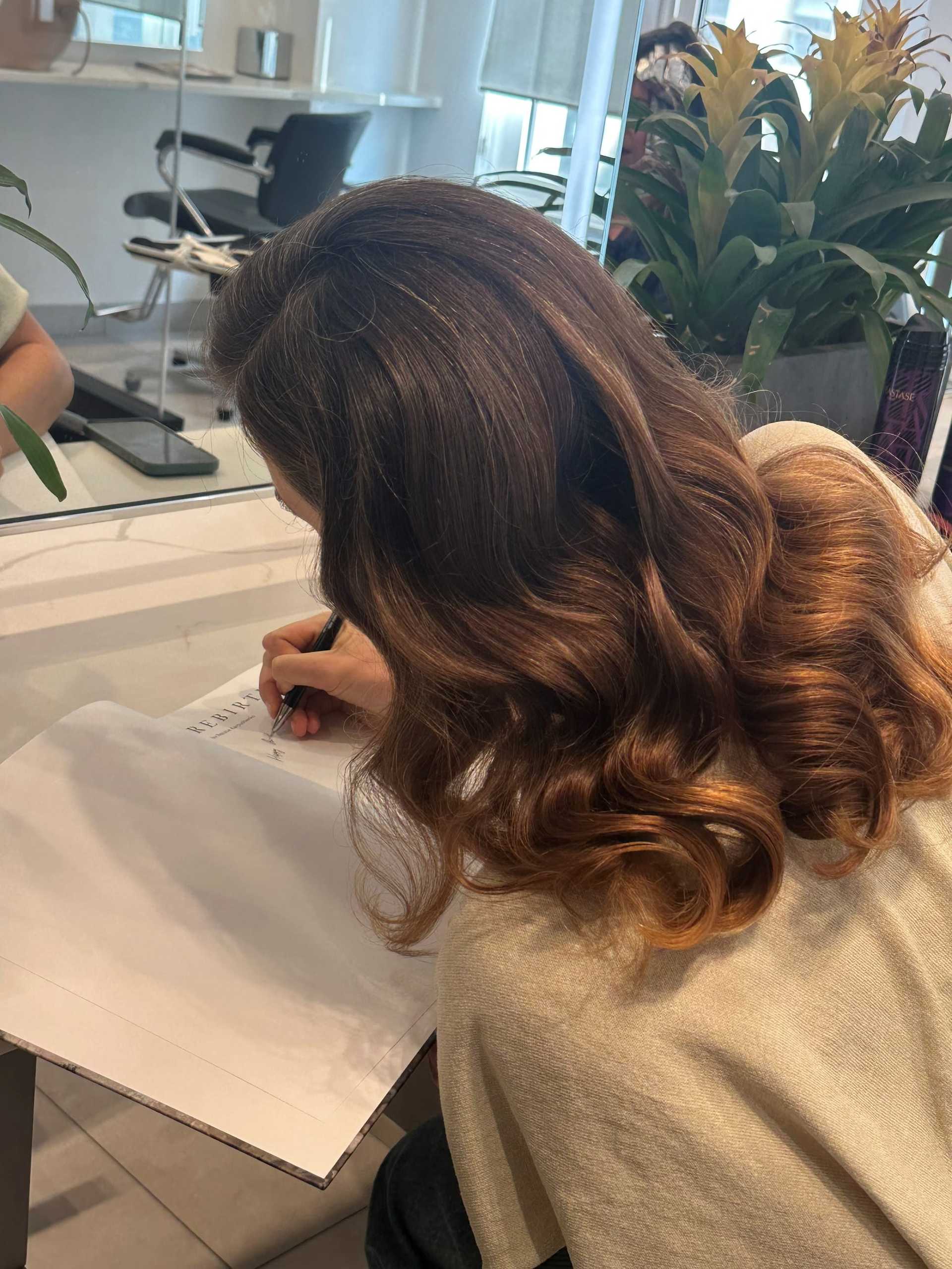 Person sitting at a table writing on a paper, with styled hair and plants in the background.