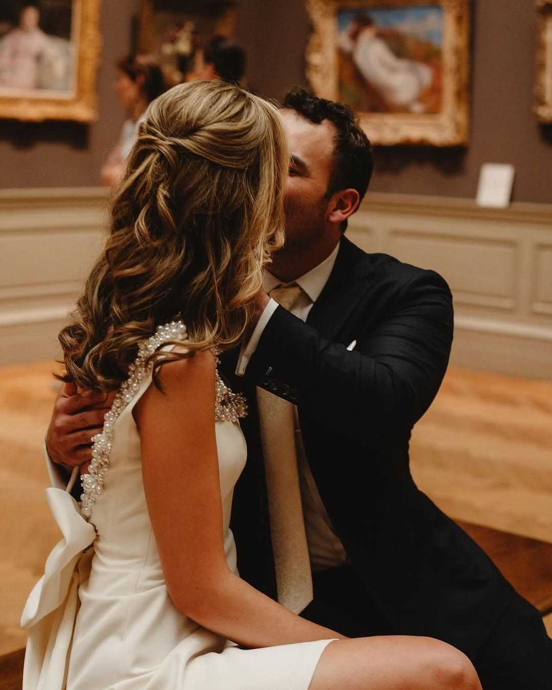 Couple embraces in art gallery, surrounded by classic paintings.