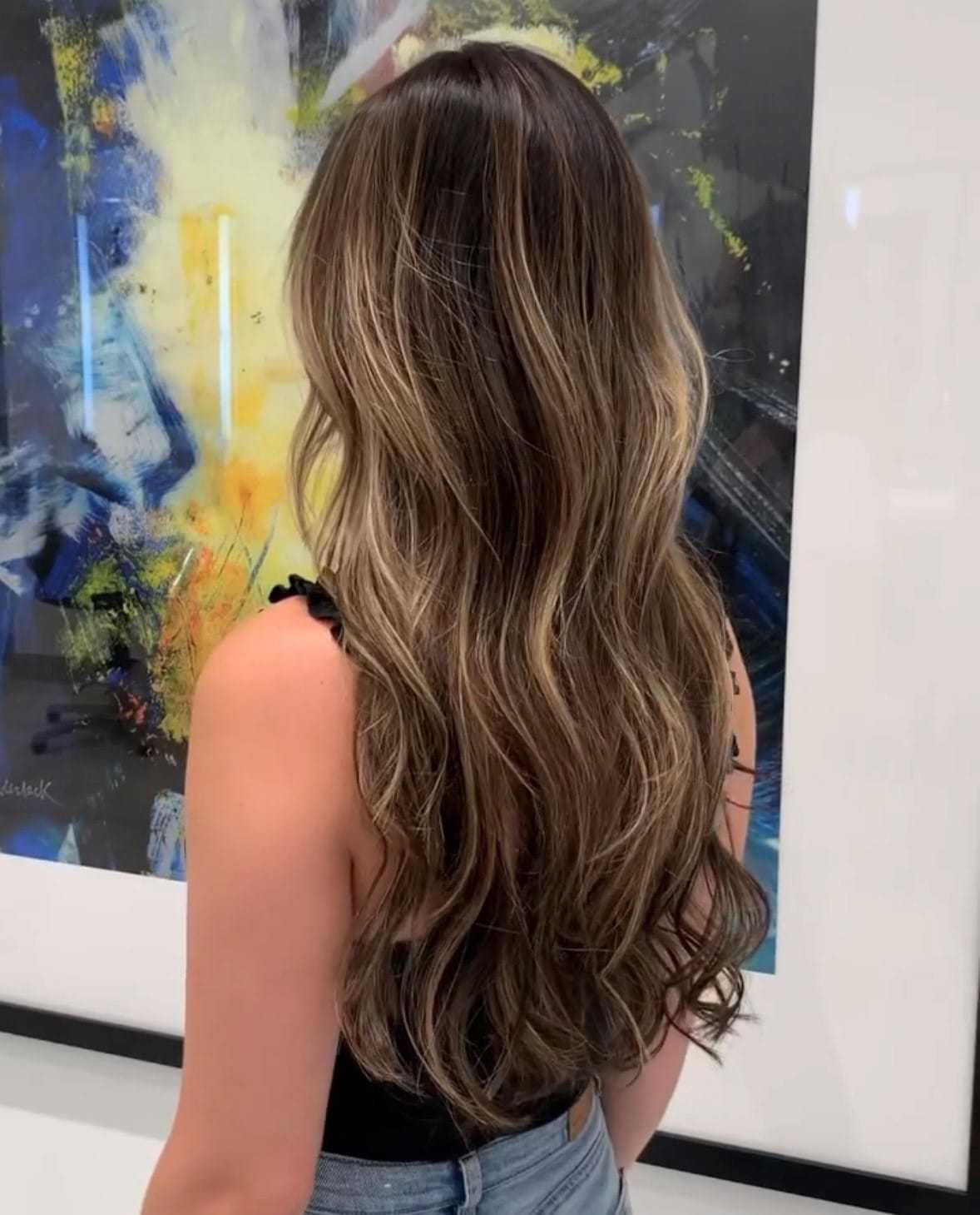 Long wavy hair with blonde highlights on a person standing in front of abstract art.